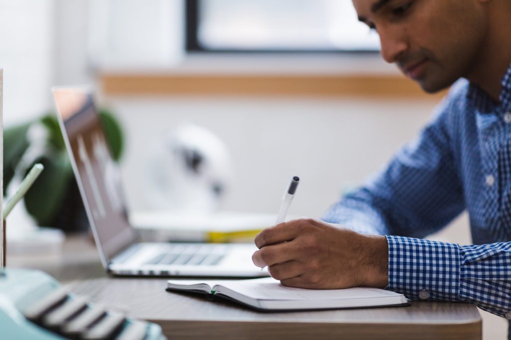 man in blue checkered shirt taking notes in front of his laptop