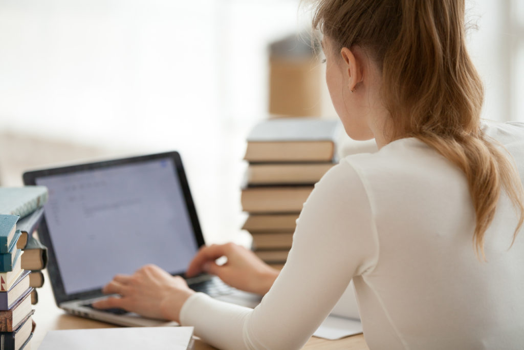 woman in a white blouse working on her laptop next to a large pile of books