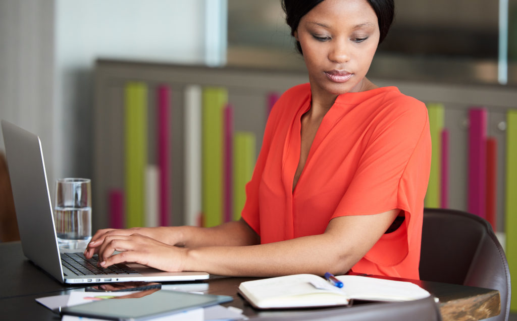 african american woman comparing notes between laptop and her notebook