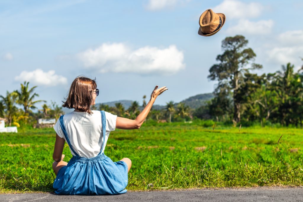 woman sitting on the ground in a rural area and throwing her hat in the sky