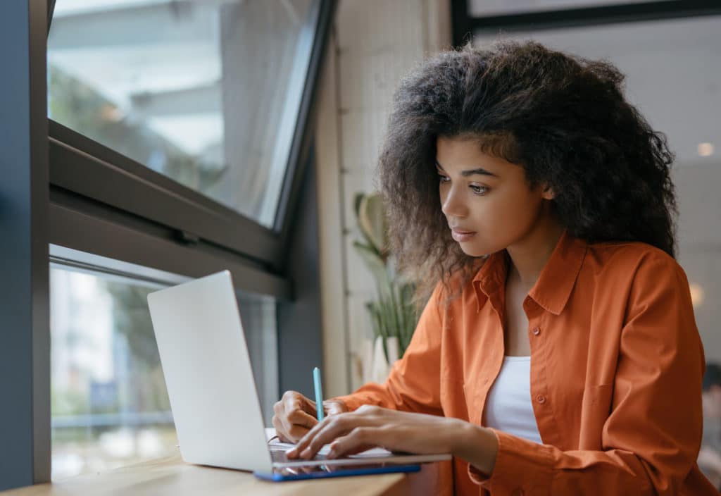 woman in a orange shirt working on her laptop next to the window