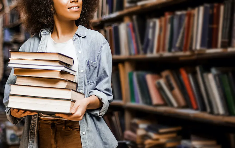 woman with a denim jacket holding a stack of books in a library