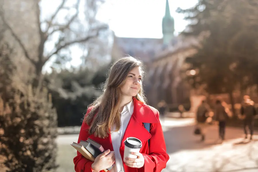 woman in a red jacket holding a cup of coffee and books in her hands