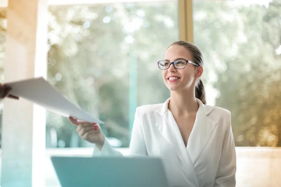 woman in a white jacket smiling while handing documents to somebody 