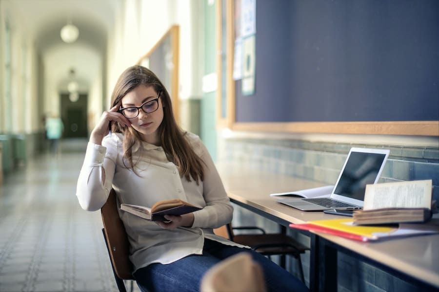 woman with eyeglasses reading a book in a library hallway