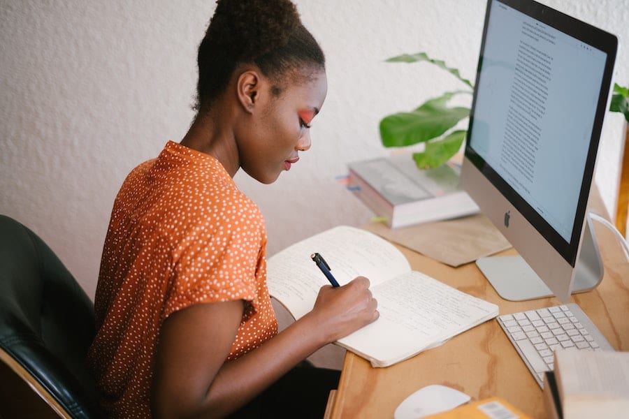 african american woman writing notes in her notebook in front of a pc
