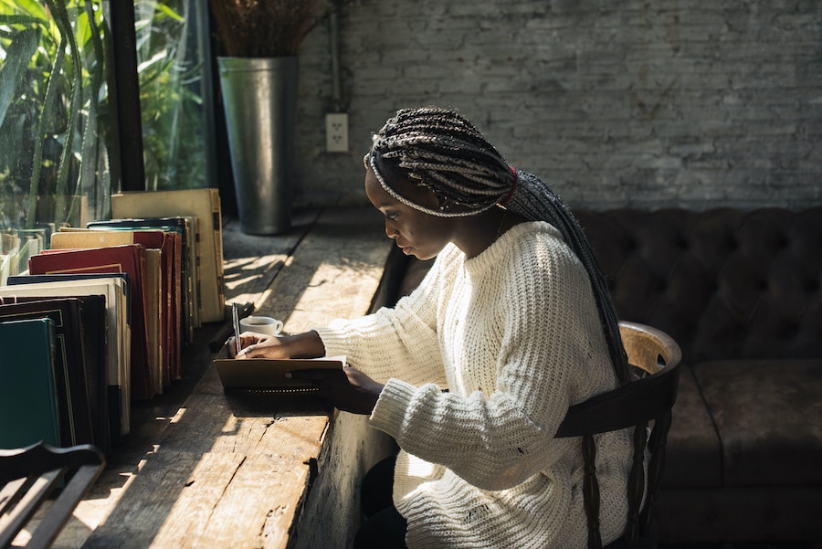 african american woman in white knitted sweater writing in a notebook by the window