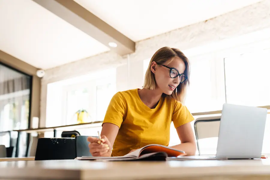 blonde woman with eyeglasses writing notes in front of her laptop