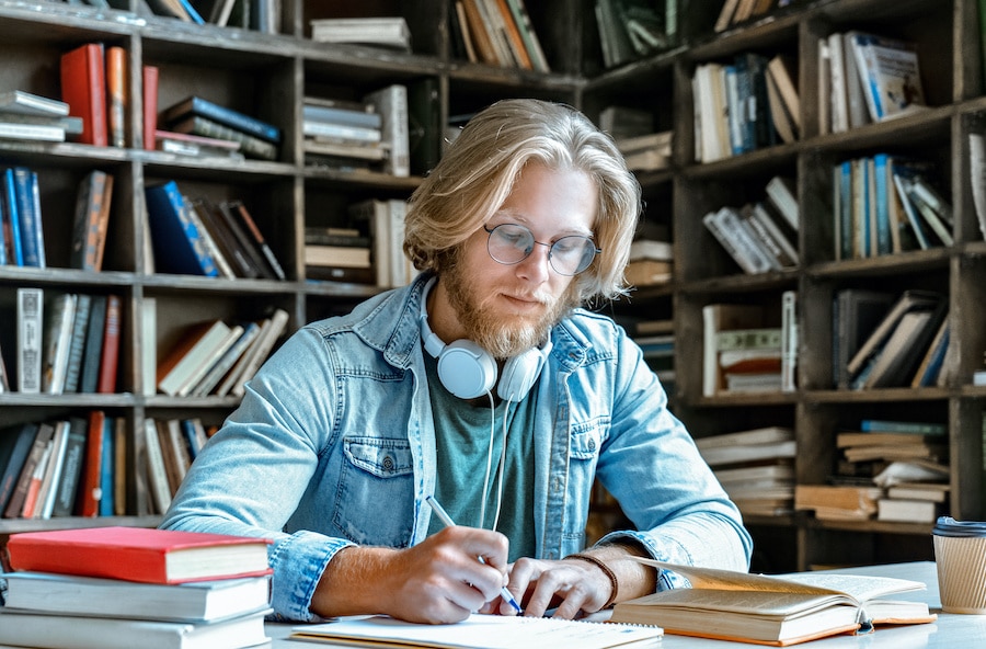 man with long blonde hair writing in a notebook in a library