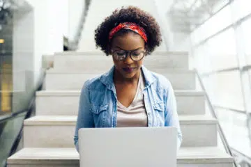 woman in a denim jacket sitting on the stairs and typing on her laptop