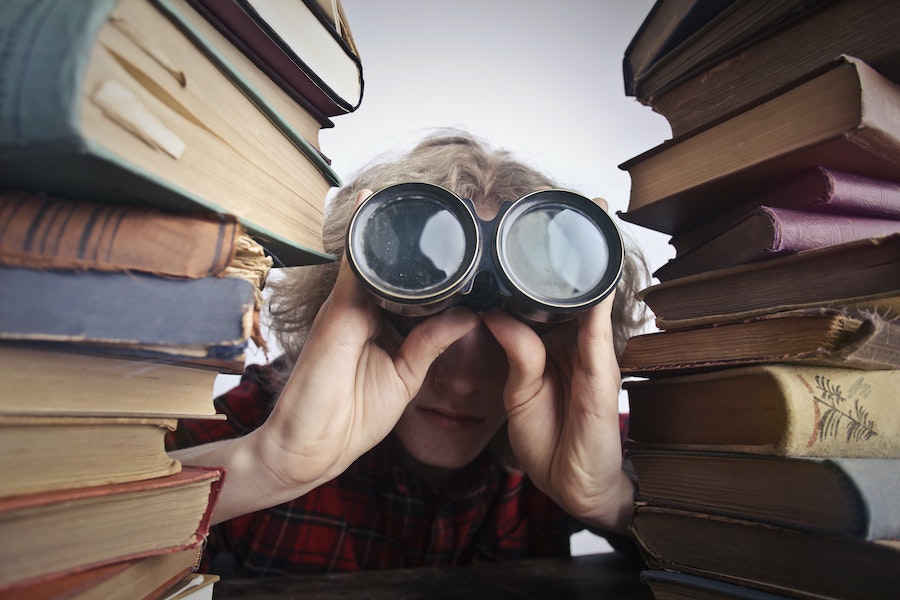 person with binoculars seeing behind a large stack of books