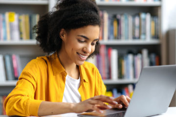 woman in a yellow jacket smilingly typing on her laptop