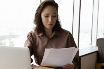 woman reading a sheet of paper in front of her laptop