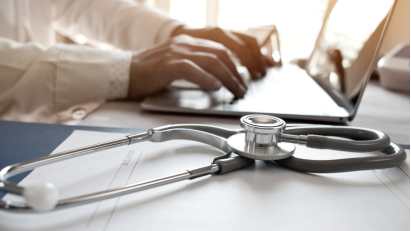 close-up shot of a stethoscope next to a doctor working on a laptop