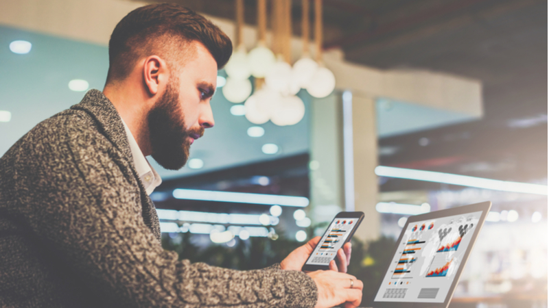 bearded man in a sweater comparing charts between phone and laptop