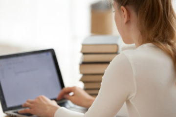 woman typing on her laptop next to a big pile of books