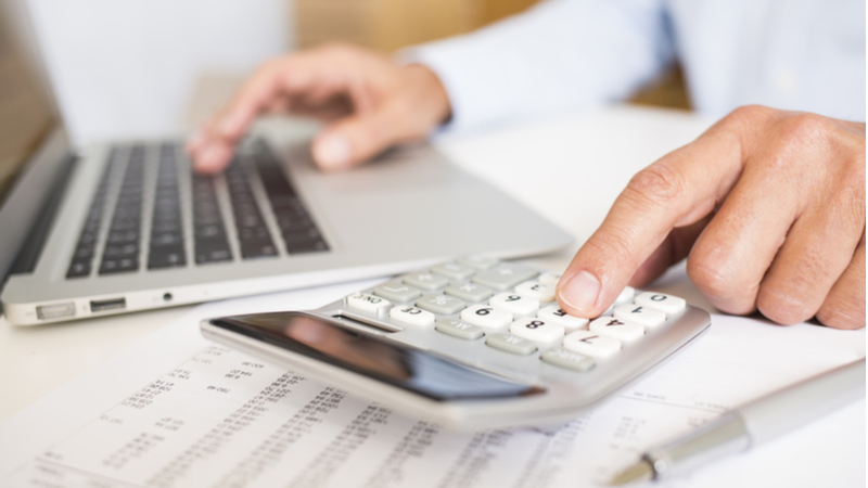 close-up man using a calculator next to his laptop