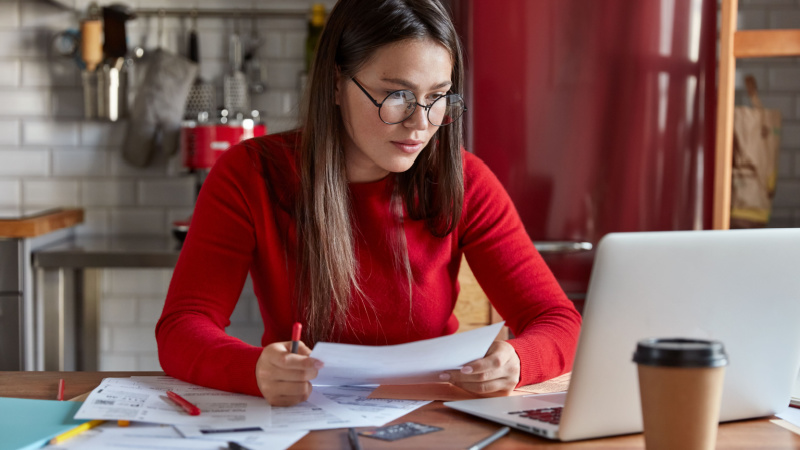 woman in red carefully analyzing documents in front of laptop